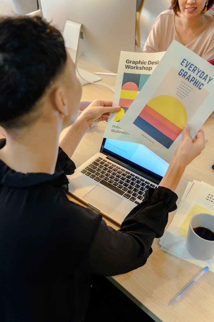 A professional reviews colorful graphic design brochures at a laptop in a modern office environment.